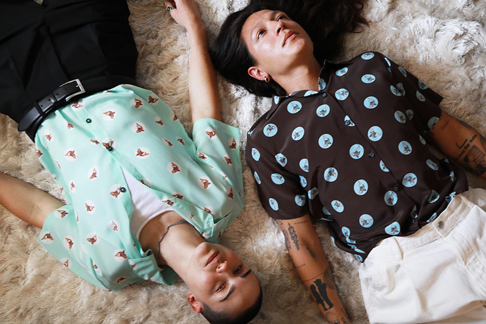 Two genderless people laying on an ivory colored rug next to each other. While wearing silk Koi fish printed shirts. One in an Aqua color and the other in an espresso color.